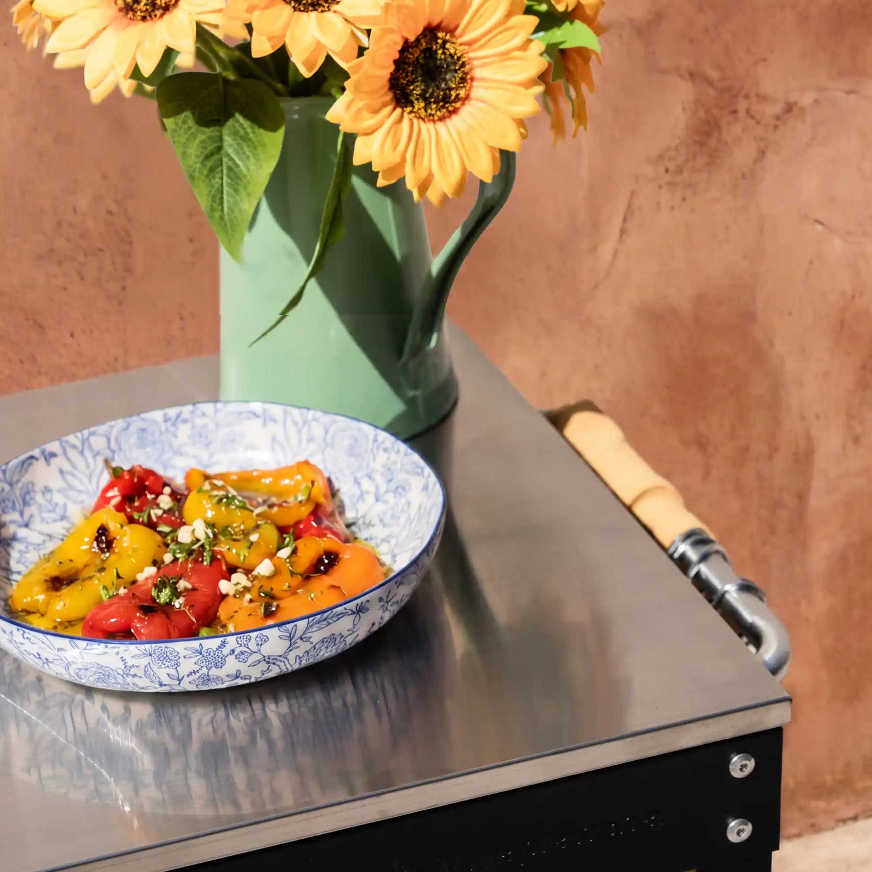 Gros plan sur le plan de travail en inox de la desserte Oliva avec bouquet de tournesols et bol de légumes grillés. ::visuel:sanscache,aveccache