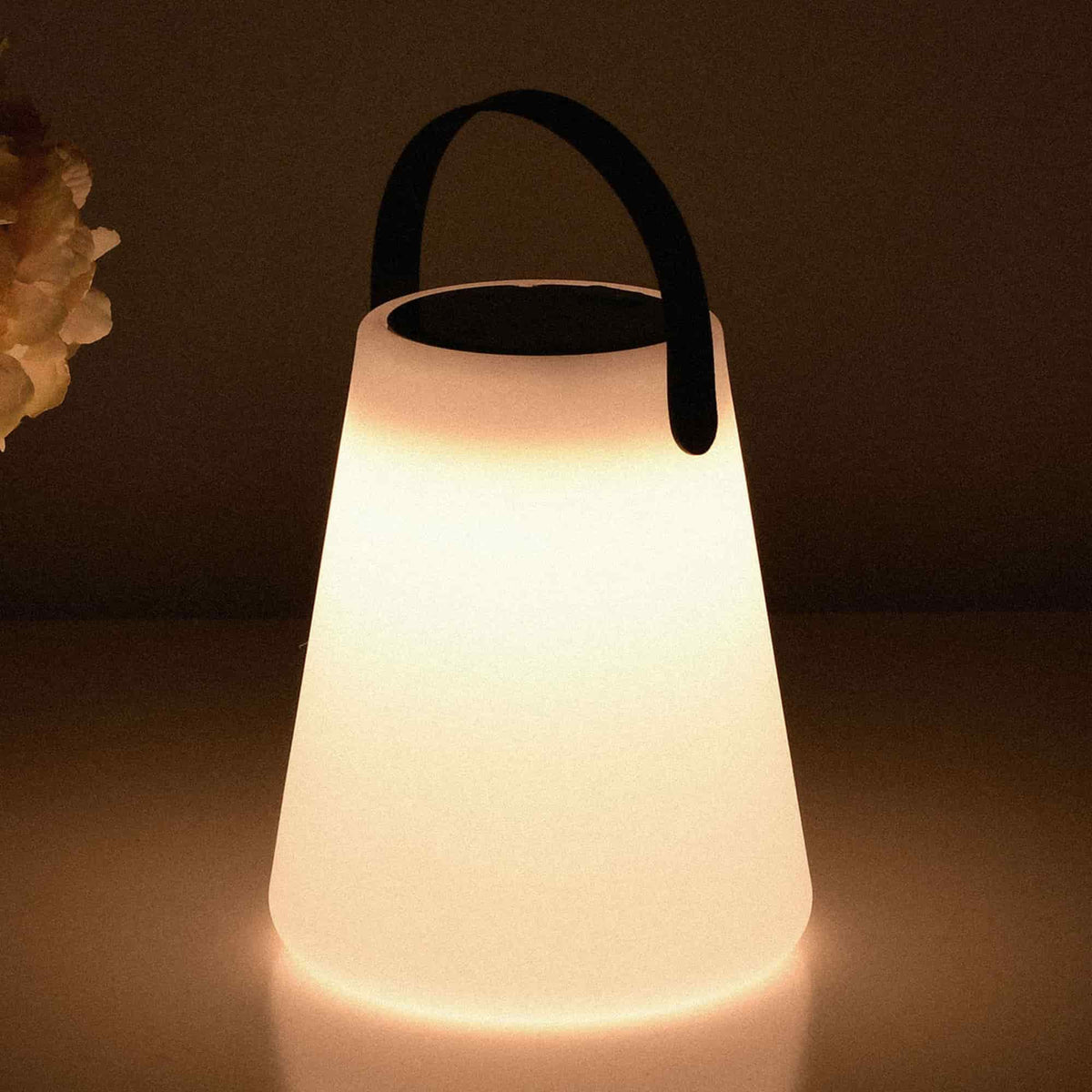 Lampe conique solaire allumée, diffusant une lumière chaude et douce, avec une anse noire en haut, posée sur une surface claire dans une ambiance tamisée.