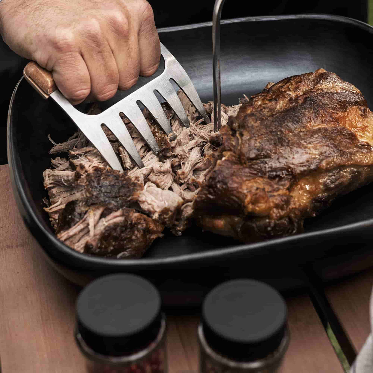 Main utilisant une griffe à viande en inox pour effilocher un morceau de viande rôti dans un plat noir, avec deux pots d’épices flous au premier plan.