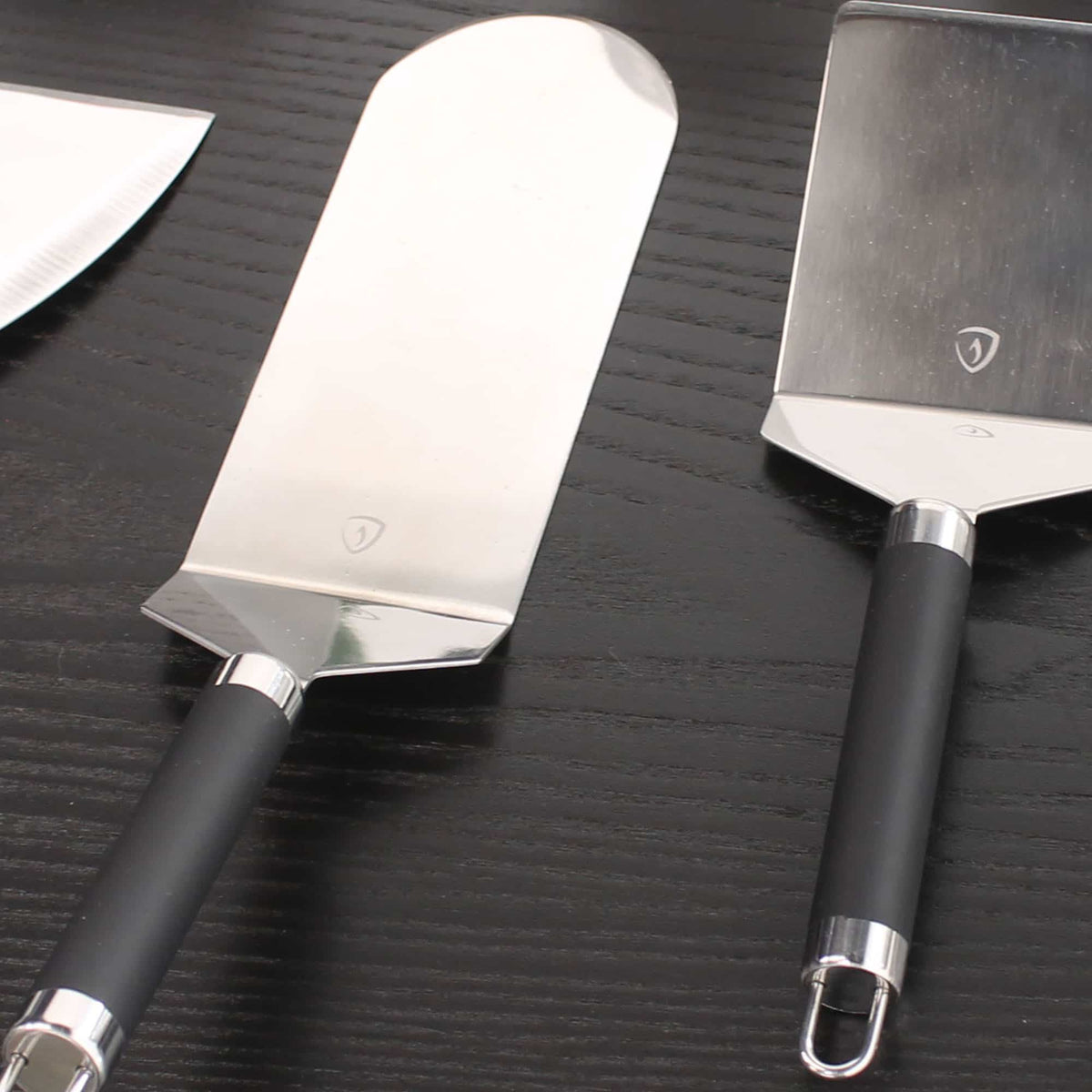 Spatules en inox à manche noir, dont une spatule longue type poisson, posées sur un plan de travail en bois foncé, montrées en gros plan.