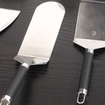 Spatules en inox à manche noir, dont une spatule longue type poisson, posées sur un plan de travail en bois foncé, montrées en gros plan.